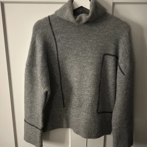 Grå stickad tröja - Mysig grå stickad tröja från Zara med hög krage och asymmetriska sömdetaljer. Tröjan har lång ärm och bred ribbad mudd vid ärmslut och nederkant. Perfekt för kyliga dagar.