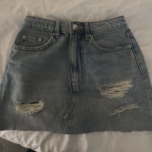 Blå jeanskjol med slitningar - Säljer en blå jeanskjol med coola slitningar framtill. Kjolen har klassiska fickor och bälteshällor, perfekt för en avslappnad stil. Medelhög midja