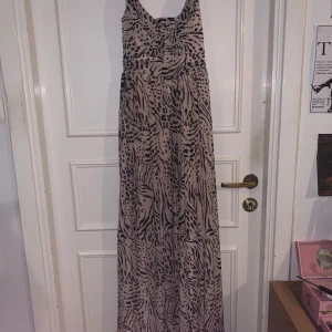 Beige långklänning med djurmönster från H&M - Säljer en beige långklänning från H&M med svart djurmönster. Klänningen har smala axelband och en figurnära överdel som övergår i ett flödande långt kjolparti. Perfekt för dig som vill sticka ut med ett coolt mönster.