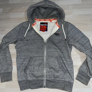 Grå zip-hoodie från Superdry - Säljer en gråmelerad hoodie från Superdry med dragkedja och huva. Tröjan har  tjocka vita snören, två fickor framtill och broderad Superdry-logga på bröstet. Perfekt för en vintage avslappnad stil. Originell storlek XL, sitter som en M-L men lutar lite mer åt L. Skriv gärna vid minsta fråga, pris kan diskuteras. Originalpris runt 799kr.
