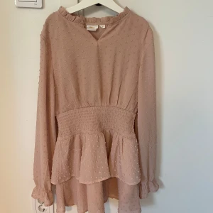 Ljusrosa klänning med volang från Gina Tricot - Supersöt ljusrosa klänning från Gina Tricot med lång ärm och volangdetaljer vid midjan. Klänningen har smockad midja och diskret prickigt mönster i tyget. Perfekt för vår och sommar!