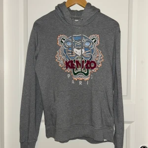 Grå hoodie med tigerbrodyr från Kenzo - Säljer en grå hoodie från Kenzo med ikonisk tigerbrodyr och texten 'KENZO PARIS' framtill. Tröjan har huva med dragsko och ribbade muddar. Tillverkad i mjuk bomull för en skön känsla.