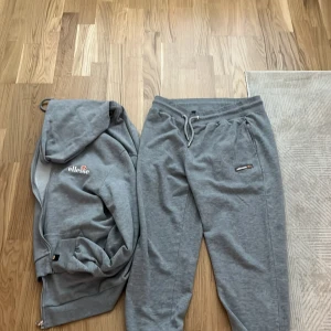 Grå mjukisbyxor från Ellesse och kofta - Gråa mjukisbyxor från Ellesse med dragsko i midjan och resår vid bensluten. Byxorna har en liten Ellesse-logga på ena benet och är tillverkade i mjukt material som passar perfekt för en avslappnad stil.