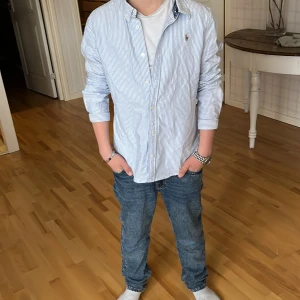 Blårandig skjorta från Ralph Lauren - Säljer en klassisk blårandig skjorta från Ralph Lauren med broderad logga på bröstet. Skjortan har långa ärmar, knappar framtill och är tillverkad i bomull. Perfekt för en stilren och avslappnad look.