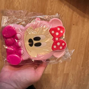 Söt rosa axelväska med musmotiv - En gullig rosa axelväska i form av en tecknad mus med stora öron och rödprickig rosett. Väskan är tillverkad i syntetmaterial och har dragkedja upptill. Perfekt accessoar för dig som gillar söta detaljer.
