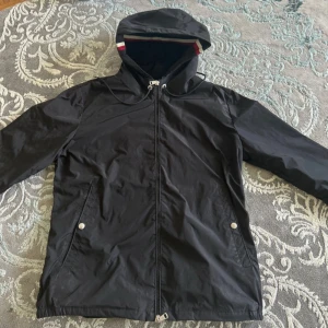 Svart vindjacka från Moncler med huva - Säljer en svart vindjacka från Moncler med huva och dragkedja framtill. Jackan har två snedställda fickor med tryckknappar och Moncler-logga på ärmen. Perfekt för blåsiga dagar och enkel att matcha.