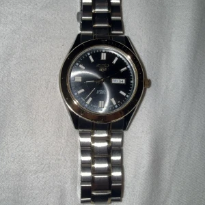 Seiko 5 automatisk herrklocka i silver och guld - Klassisk Seiko 5 automatisk klocka med svart urtavla, dag- och datumvisning samt silverfärgad länk med gulddetaljer. Urtavlan har tydliga index och visare. Baksidan visar den automatiska mekanismen.