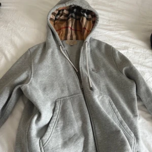 Grå hoodie från Burberry med klassiskt rutigt foder - Säljer en grå hoodie från Burberry med dragkedja och kängurufickor. Insidan av huvan har det ikoniska Burberry-rutiga fodret i beige, rött och svart. Perfekt för en avslappnad och stilren look.