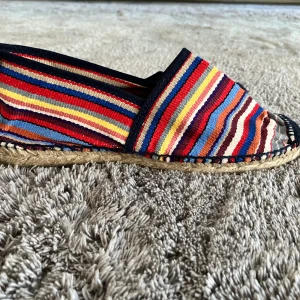 Färgglada randiga espadrillos från Toni Pons - Säljer ett par espadrillos från Toni Pons med färgglada ränder i rött, blått, gult och orange. Skorna har ovandel i textil och sula i flätad jute och yttersula i gummi. Perfekta för varma dagar och ger en bohemisk touch till outfiten. 