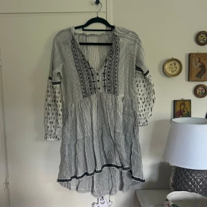 Vit och svart mönstrad klänning med broderi - Säljer en luftig vit klänning med svarta broderade detaljer och v-ringning. Klänningen har långa ärmar, knappar framtill och sidofickor. Modellen är lös och bekväm, passar perfekt som en sommarklänning eller strand cover-up🥰