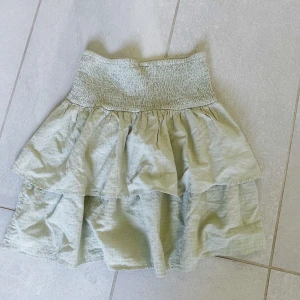 volangkjol  - Säljer en söt grön kjol med dubbla volanger och smockad midja. Kjolen har en luftig känsla och passar perfekt till varma dagar. Enkel att matcha med olika toppar för en avslappnad stil.