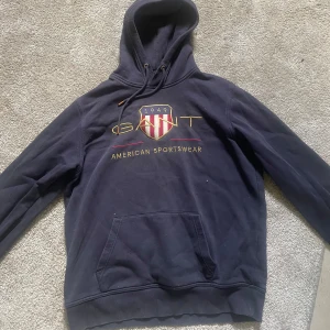 Mörkblå hoodie från GANT - Säljer en mörkblå hoodie från GANT med broderad logga och texten 'American Sportswear' på bröstet. Tröjan har huva med dragsko och en stor magficka. Perfekt för en sportig och avslappnad stil.