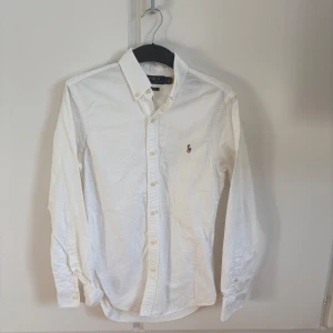 Vit skjorta från Polo Ralph Lauren - Klassisk vit skjorta från Polo Ralph Lauren med broderad logga på bröstet. Skjortan har knappar framtill och krage, samt långa ärmar. Perfekt till många olika tillfällen. 