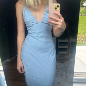 Ljusblå långklänning med öppen rygg och v-ringning - Säljer en ljusblå långklänning med smala axelband, djup v-ringning och öppen rygg med knyt. Klänningen har en figurnära passform och är perfekt för sommarens tillfällen. Materialet känns lätt och svalt.