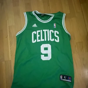 Grön Boston Celtics baskettröja Rondo #9 - Säljer en grön Boston Celtics baskettröja storlek L-XL från Adidas med vitt tryck, nummer 9 och namnet Rondo på ryggen. Tröjan har vita detaljer vid ärmslut och hals, samt officiell NBA Team Apparel-märkning. Perfekt för basketfans! 