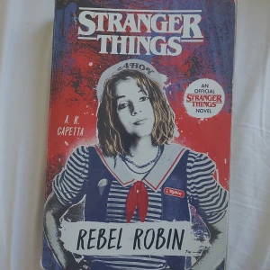 Stranger Things: Rebel Robin - Säljer boken 'Stranger Things: Rebel Robin' av A.R. Capetta. En officiell Stranger Things-roman med färgstarkt omslag i rött, blått och vitt, med en bild på Robin i skoluniform. Perfekt för fans av serien och ungdomsböcker.