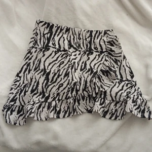 Zebramönstrad volangkjol från 157 - Säljer en kort zebramönstrad kjol från 157 med volangkant. Kjolen har svartvitt mönster och bred resår i midjan för en bekväm passform. Perfekt för dig som gillar djurmönster och vill sticka ut lite extra.