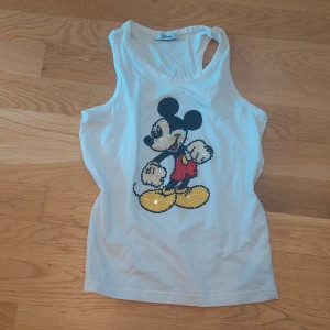 Vit linnetopp med paljett-Musse Pigg från Disney Signature - Vit ärmlös linnetopp med stort paljettmotiv av Musse Pigg på framsidan. Perfekt för dig som gillar Disney och vill ha något lekfullt och glittrigt i garderoben.