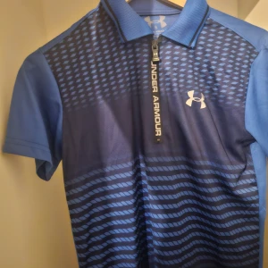 Blå pikétröja från Under Armour - Säljer en blå pikétröja från Under Armour med kort dragkedja och mönstrad ovandel. Tröjan har korta ärmar och är tillverkad i ett syntetmaterial som andas. Perfekt för träning eller sportiga aktiviteter.
