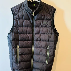Svart dunväst från Hugo Boss - Snygg svart  dunväst från Hugo Boss med dragkedja framtill och två fickor med dragkedja. Västen har en hög krage och diskret logotyp på bröstet. Perfekt för lager-på-lager under kyliga dagar.