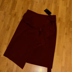 Vinröd omlottkjol med knytband - Vinröd omlottkjol från Selected Femme med knytband i midjan. Kjolen har en enkel och stilren design med rak passform och slät yta. Perfekt för dig som gillar minimalistisk stil och vill ha en kjol som sticker ut med sin färg.