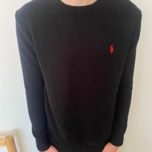Svart stickad tröja från Ralph Lauren - Klassisk svart stickad tröja från Ralph Lauren med rund halsringning och röd broderad logga på bröstet. Tröjan har långa, smala ärmar. Perfekt för en stilren och tidlös look. Skriv vid minsta funderingar!