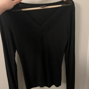 Svart långärmad topp med spetsdetalj - Svart långärmad topp från Gina med v-ringning och fina spetsdetaljer längs halsringningen i storlek S. Har enbart använt en gång. Nypris 280.