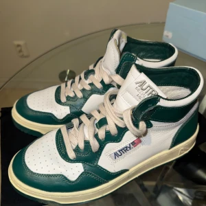 Autry Medalist Mid vita och gröna sneakers - Säljer ett par Autry Medalist Mid sneakers i vitt och grönt. Skorna har högt skaft, snörning och perforerad tå för extra ventilation. Klassisk retrostil med märkets logga och amerikanska flaggan på sidan. Perfekta för dig som gillar streetwear.