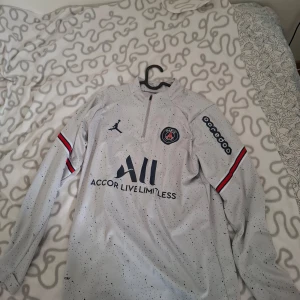 PSG långärmad fotbollströja från Jordan - Säljer en vit långärmad PSG fotbollströja från Jordan med svarta och blå prickar, klubbmärke på bröstet och sponsorloggor. Tröjan har dragkedja vid halsen och röda/blå detaljer på ärmarna. Storlek herr M.