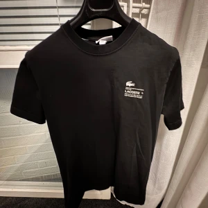 Svart t-shirt från Lacoste - Säljer en svart t-shirt från Lacoste med vit logga och text på bröstet. Klassisk rund halsringning och korta ärmar. Perfekt för en clean och stilren look.