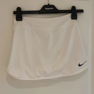 Vit tenniskjol från Nike - Säljer en vit tenniskjol från Nike med inbyggda shorts under. Kjolen har en klassisk och sportig design med Nike-logga på nederkanten. Perfekt för dig som gillar sportig stil.