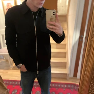 Svart overshirt med dragkedja - Säljer en stilren svart overshirt med hel dragkedja framtill och klassisk krage. Jackan har två stora bröstfickor och lång ärm, perfekt för lager-på-lager-stil. Passar till många olika outfits.