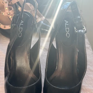 Svarta lackpumps från ALDO med rem - Säljer ett par svarta pumps från ALDO i glansigt lackmaterial. Skorna har en smal rem runt hälen och hög klack. Perfekta för dig som vill ha en klassisk och elegant look.