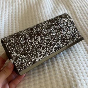 Glittrig svart kuvertväska från La Regale - Säljer en svart kuvertväska från La Regale med glittrig framsida och metallkant. Väskan har en kedja som axelrem och stängs med magnetlås. Perfekt bal/fest eller  för att addera lite extra bling till din outfit.