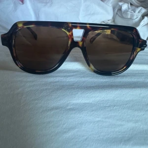 Bruna solglasögon med leopardmönster - Snygga solglasögon med bred båge i brunt leopardmönster och mörka glas. Modellen har en unik detalj vid näsryggen och ger en cool oversized look. Perfekt accessoar för att lyfta din stil.