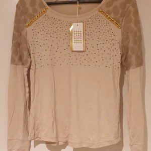 Beige långärmad topp med kedjedetaljer och nitar från Landmark - Säljer en beige långärmad topp från Landmark med transparenta prickiga partier på axlar och ärmar. Framsidan är dekorerad med små nitar och guldfärgade kedjor vid axlarna. Perfekt för dig som vill sticka ut med snygga detaljer.