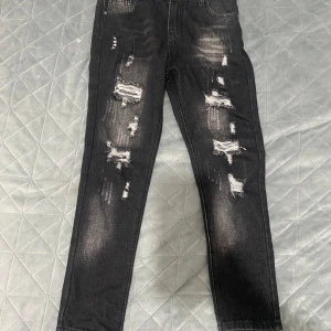 Svarta slitna jeans med raka ben - Säljer ett par svarta jeans med tydliga slitningar och hål på både fram- och baksida. Jeansen har raka ben och klassisk femficksdesign. Perfekta för dig som gillar en edgy och trendig stil.