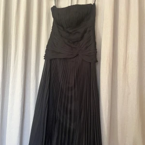 Svart balklänning med plisserad kjol - Elegant svart långklänning med axelbandslös överdel och draperade detaljer framtill. Kjolen är plisserad vilket ger ett snyggt fall. Perfekt för bal eller fest. Klänningen har en klassisk och tidlös design. Det är en S men passar även M.