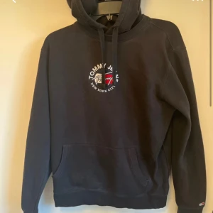 Svart hoodie från Tommy Hilfiger - Svart hoodie från Tommy Hilfiger med broderad logga och texten 'New York City' på bröstet. Klassisk känguruficka framtill och ribbade muddar. Perfekt för en avslappnad och stilren look. ANVÄND EN GÅNG, nypris = 1100 kr mitt= 249 kr, skriv om minsta lilla fundering!