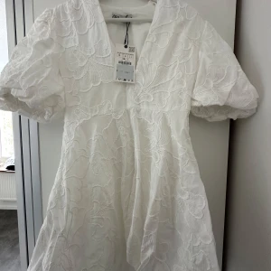 Vit kortklänning/Studentklänning från Zara - Superfin vit klänning från Zara med stora blommiga broderier och v-ringning. Klänningen har korta puffärmar och en lätt utställd kjol. Perfekt för sommarens alla tillfällen!