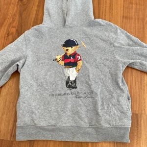 Grå hoodie med Polo Bear från Ralph Lauren - Säljer en grå hoodie från Ralph Lauren med den klassiska Polo Bear-trycket på framsidan. Tröjan har huva och är tillverkad i mjukt material, perfekt för en avslappnad stil. Ikonisk och tidlös design! Tröjan passar de som är 150-161cm