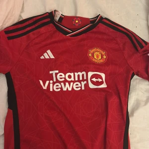 Manchester United hemmatröja Garnacho 17 - Röd Manchester United fotbollströja med Garnacho 17 på ryggen. Adidas-logga på bröstet, klubbmärke och sponsortryck framtill. Svarta detaljer på axlarna och vita tryck. Klassisk rund hals och korta ärmar.storlek 152, 11-12 år