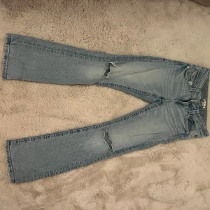 Ljusblå bootcut jeans med slitningar - Säljer ett par ljusblå jeans med bootcut och är low waisted i passformen. De har slitningar på båda benen. Jeansen har klassiska fem fickor och dekorativa knappar på bakfickorna. Perfekta för en avslappnad stil. Har ett litet slitage mellan bakfickorna.