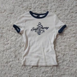 Vit t-shirt marinblå detaljer - Supersörit t-shirt med marinblå kant vid hals och ärmslut. Tryck på bröstet med texten 'Sail Away with me' och ett ankare. Passformen är ganska tight med korta ärmar. Perfekt till sommaren eller för dig som söker en 'coastal girl' look!
