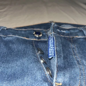 Blå Dsquared2 jeans med tryck och slitningar - Säljer ett par blå jeans från Dsquared2 med vita tryckdetaljer både fram och bak. Jeansen har slitningar och hål på benen, knappar i gylfen och klassisk femficksdesign. Modellen är rak och har normal passform.