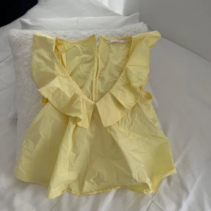 Gul volangtopp med v-ringning - Säljer en söt gul topp med djup v-ringning och stora volangdetaljer längs axlar och halsringning. Perfekt för vår och sommar, ger en fräsch och lekfull look.