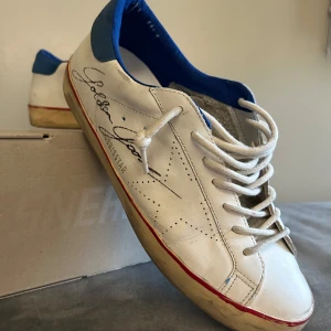 Golden Goose sneakers - Golden Goose Superstar sneakers med blå häl och röd rand längs sulan. Klassisk design med snörning och perforerade detaljer på sidorna. Märkets logga syns på sidan. Perfekta för dig som gillar en unik och trendig look. Allt följer med som jag fick vid köpet i butik (Box,kvitto dustbag samt påse) Det är bara att fråga om ni vill ha mer bilder eller några andra frågor! Köpta av mig i butik i Spanien.
