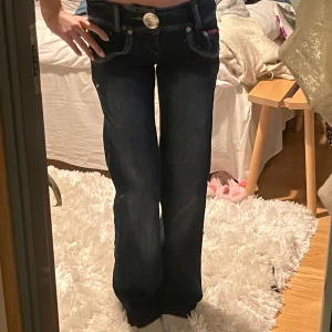 Mörkblå vida  jeans från Sister Point - Mörkblå jeans från Sister Point med vida ben och låg midja. Snygga kontrastsömmar, stora fickor bak och en tydlig logga på insidan av linningen. Med stor silverfärgad knapp framtill.