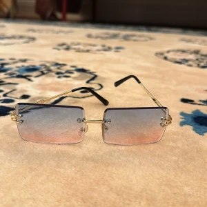 Rektangulära solglasögon med guldiga bågar och tonade glas - Snygga solglasögon med rektangulära, kantiga linser som är tonade i grått och rosa. Glasögonen har tunna guldiga metallbågar och svarta skalmar. Perfekt accessoar för en trendig look. Fodral tillkommer