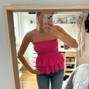 Rosa tubtopp med volang från Hollister - Säljer en rosa tubtopp från Hollister med smockad överdel och dubbla volanger nedtill. Toppen är axelbandslös och har en snygg, croppad passform. 